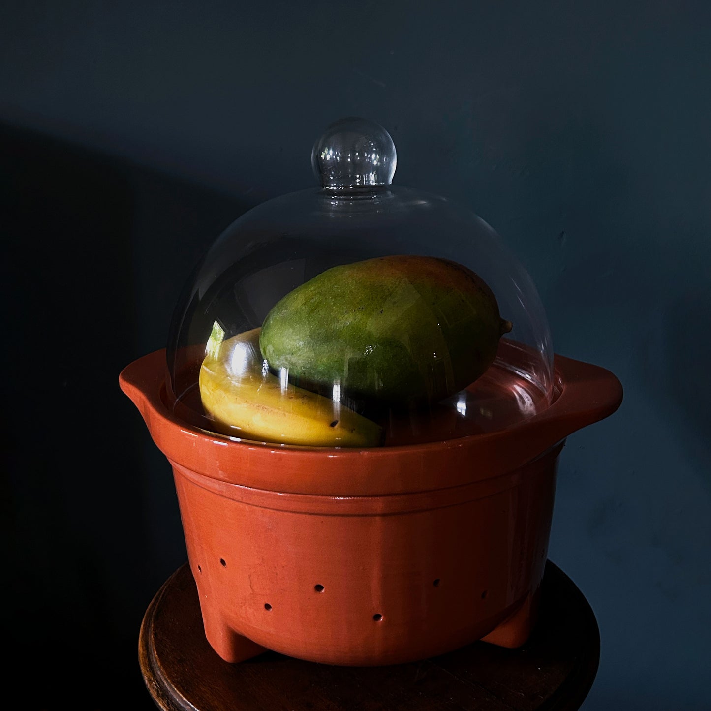 A terracotta ceramic fruit basket with a clear glass dome lid, containing a green mango and a yellow banana, set against a dark blue background.