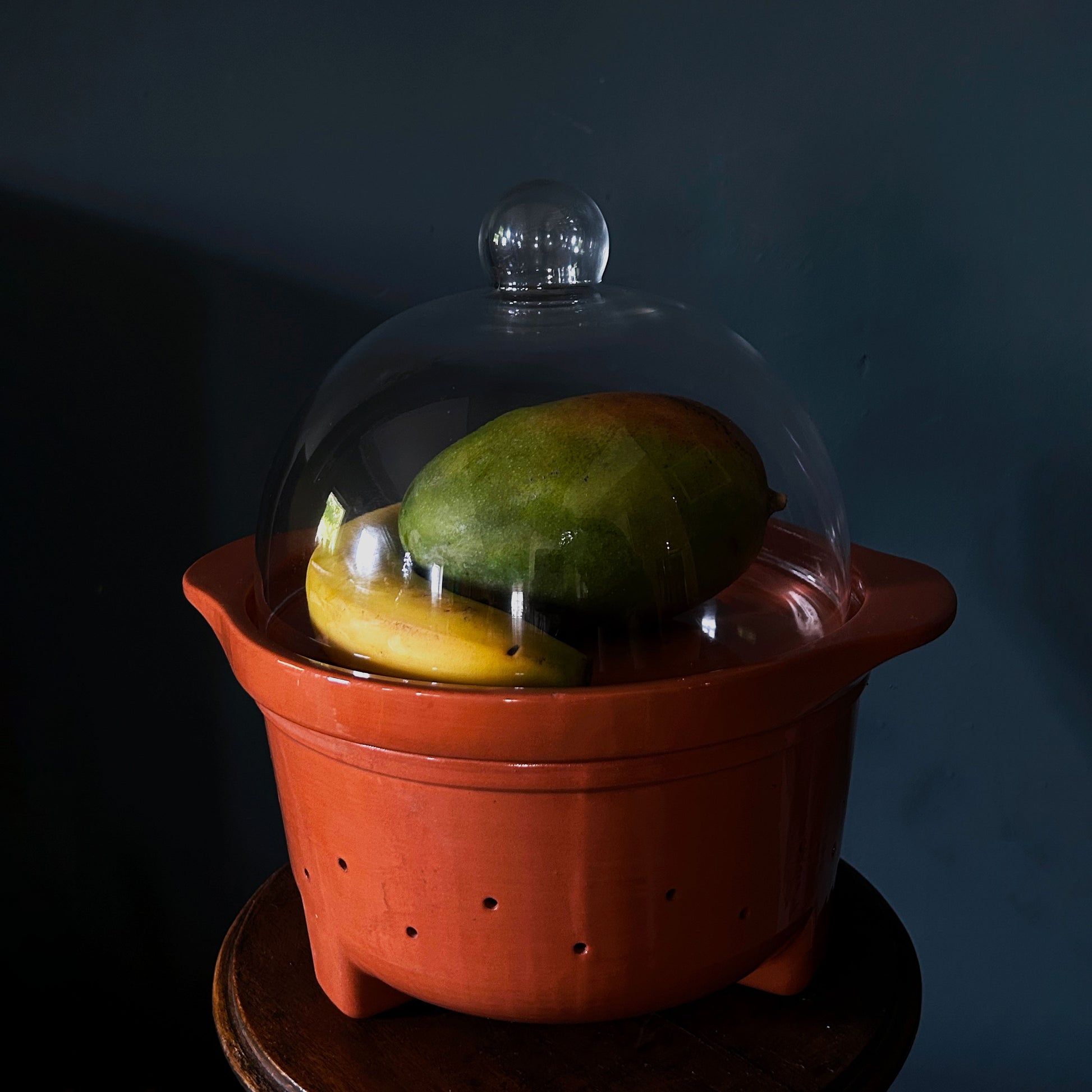 A terracotta ceramic fruit basket with a clear glass dome lid, containing a green mango and a yellow banana, set against a dark blue background.