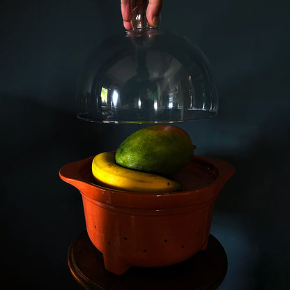 A terracotta ceramic fruit bowl with a clear glass cloche lid being lifted by a hand, containing a green mango and a yellow banana, set against a dark background.