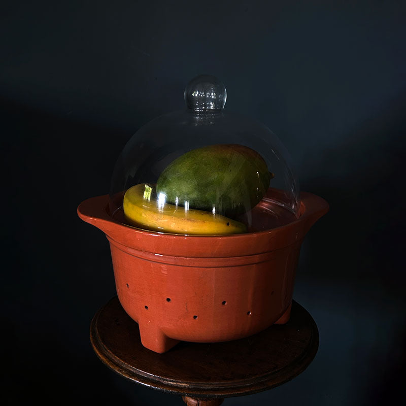 A terracotta ceramic pot with a clear glass dome lid, containing a green mango and a yellow banana, displayed on a dark wooden pedestal against a dark background.