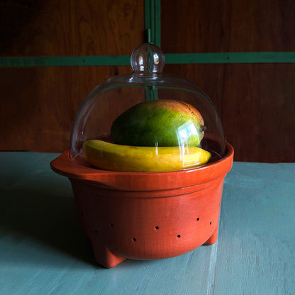 A terracotta fruit bowl with a clear glass dome lid, containing a yellow banana and a green mango, set on a light blue wooden table.