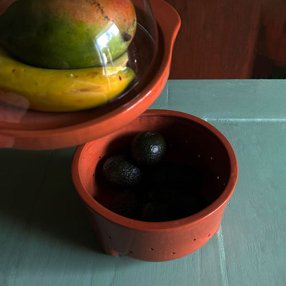 A terracotta fruit bowl shown in use, with a lid holding a mango and banana above an open basket containing dark avocados, set on a painted teal tabletop.