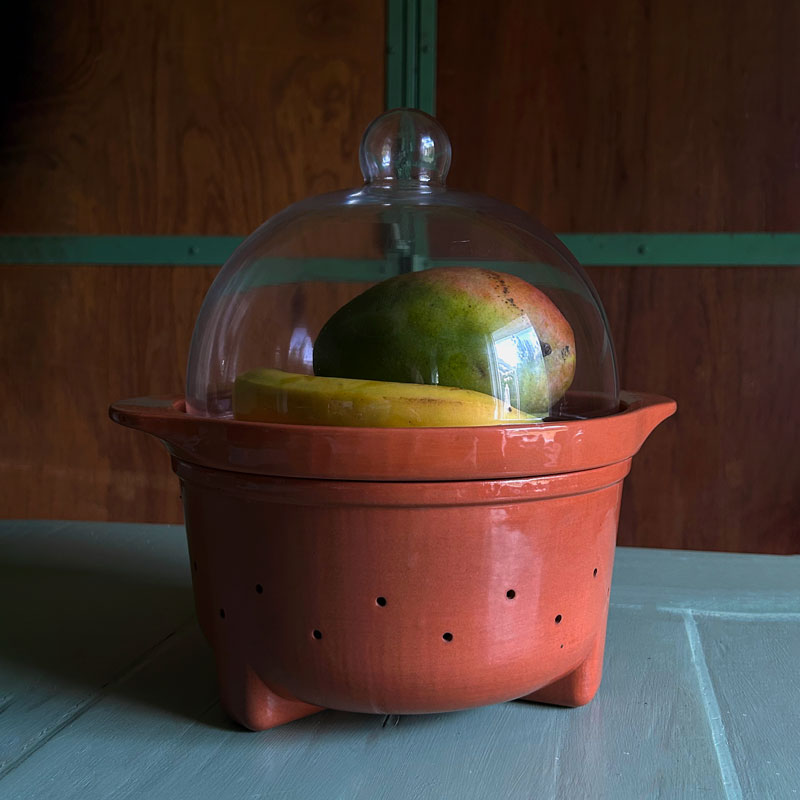 A terracotta fruit ripening bowl with a clear glass dome lid, containing a mango and a yellow fruit, set against a dark wooden background.