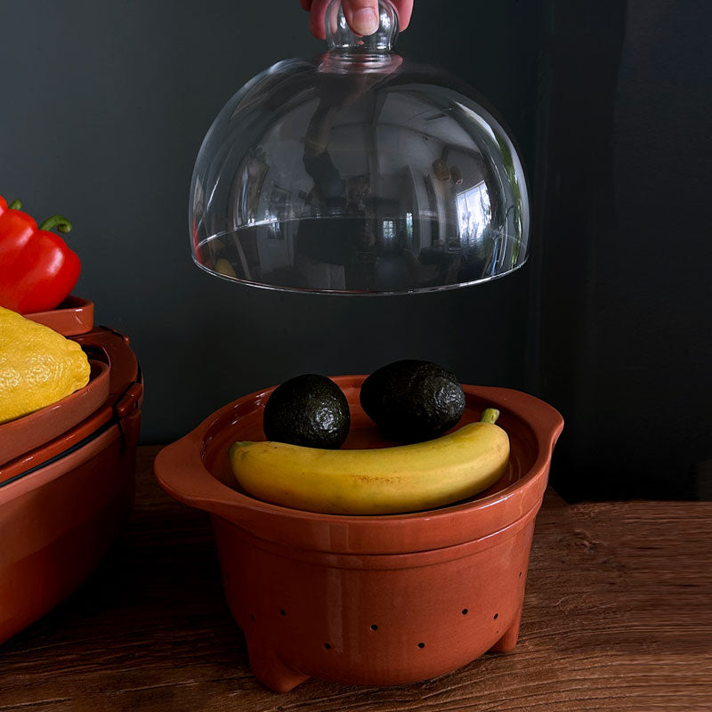 A terracotta-colored ceramic fruit and vegetable storage dish with a matching glass cloche lid being lifted by a hand, shown on a wooden table with bananas, avocados, and other vegetables.