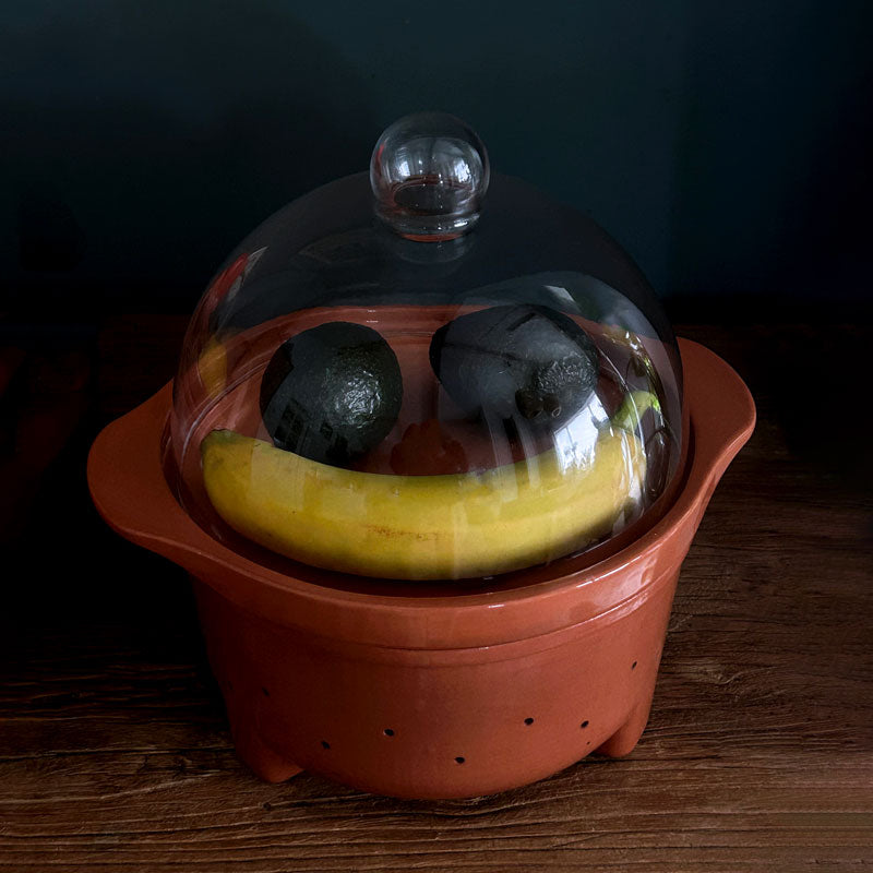 A terracotta fruit bowl with a clear glass dome lid, shown on a dark wooden surface with a banana and two avocado inside.