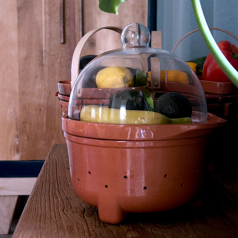 A terracotta ceramic fruit ripening bowl with a clear glass dome lid, shown filled with produce on a wooden table.