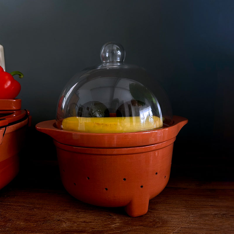A terracotta ceramic fruit ripening bowl with a clear glass dome lid, shown on a wooden surface against a dark background.